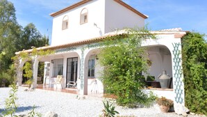 A white building with a tiled roof, a covered patio area with chairs, and a garden with plants and a large pot. - Lake Vinuela peaceful holiday in beautiful surroundings  (Los Romanes)