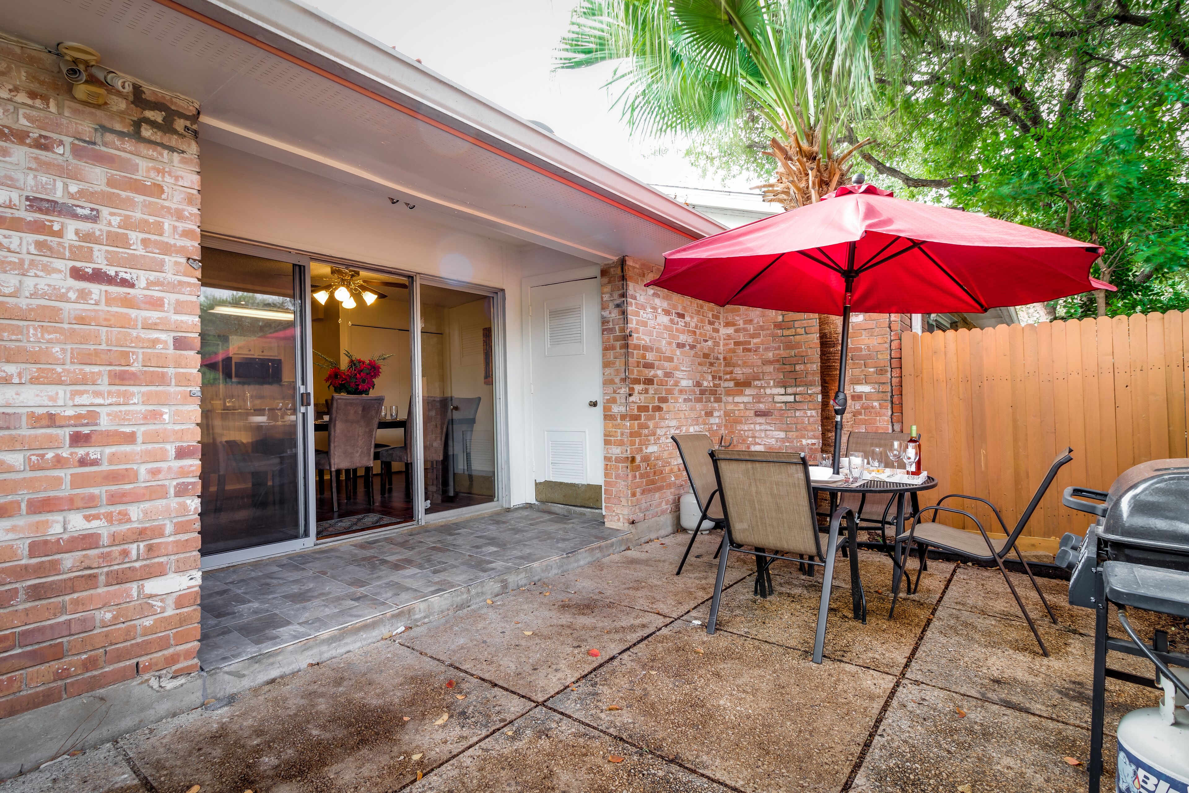 Eine Terrasse mit Ziegelmauer, einer Glasschiebetür zu einem Innenbereich, einem roten Sonnenschirm, einem Tisch mit Stühlen und einem Grill.
