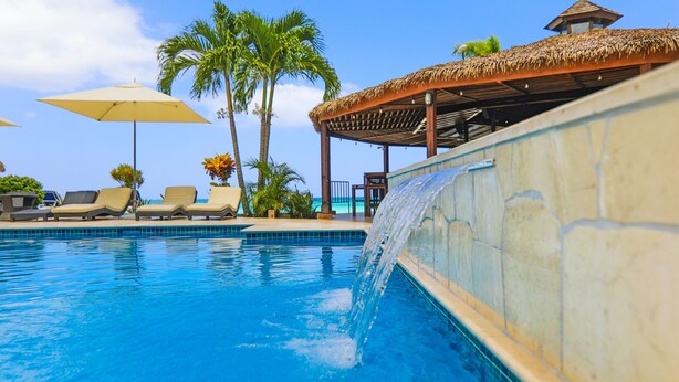 Una piscina con sillas de descanso y sombrillas, una cascada y una estructura con techo de paja.