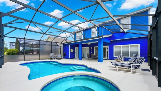 A covered outdoor swimming pool with blue and white lounge chairs, a ceiling fan, and a view of a blue house in the background. - 5 STAR REVIEWED SOUTH FACING LUXURY GOLF AND DISNEY VILLA ON GATED COMMUNITY (Davenport)