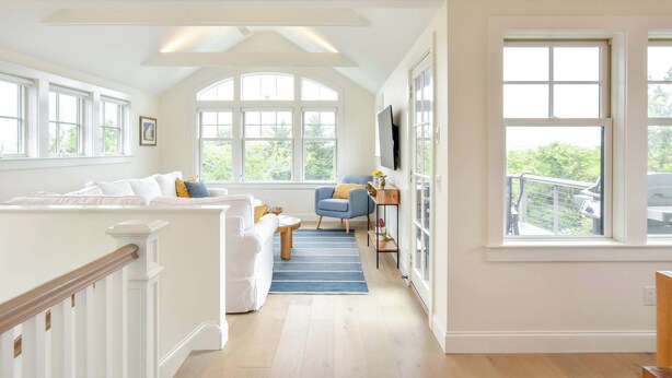 Uma sala de estar ampla com um sofá branco, uma poltrona azul e uma mesa de centro de madeira. Grandes janelas proporcionam luz natural e vista para o verde lá fora.
