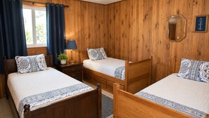 A room with wooden panel walls, two beds with matching headboards, a ceiling fan, a window with curtains, and a small table with a lamp.
