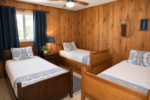A room with wooden panel walls, two beds with matching headboards, a ceiling fan, a window with curtains, and a small table with a lamp. - Z House | Shaded Coastal Hideaway on the dunes (Dauphin Island)