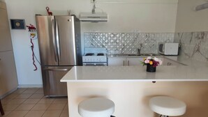 A modern kitchen with a stainless steel refrigerator, a gas stove, and a microwave. There are two white bar stools and a small potted plant on the counter. - Sanctuary for family and friends (Balcatta)