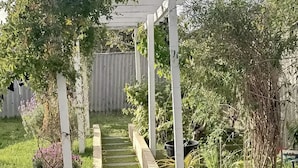 A garden pathway leading to a white pergola structure surrounded by lush greenery and a concrete path. - Sanctuary for family and friends (Balcatta)
