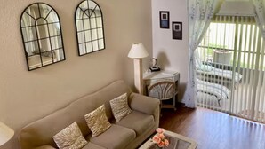 A living room with a beige sofa, a glass coffee table with flowers, a lamp, and two wall-mounted mirrors. - Unique style 2 BR/2 BA townhouse in Disney Area (Kissimmee)