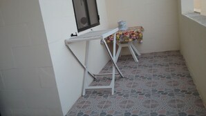 A small, enclosed balcony with a tiled floor, a white table with a floral tablecloth, and a window with a screen.