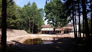 A two-story house with a deck, surrounded by a forest and a small pond.