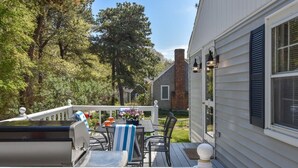 A deck with a grill, outdoor table, and chairs, surrounded by trees and a house. - Lovely Home Minutes from Beach in Chatham! (Chatham)