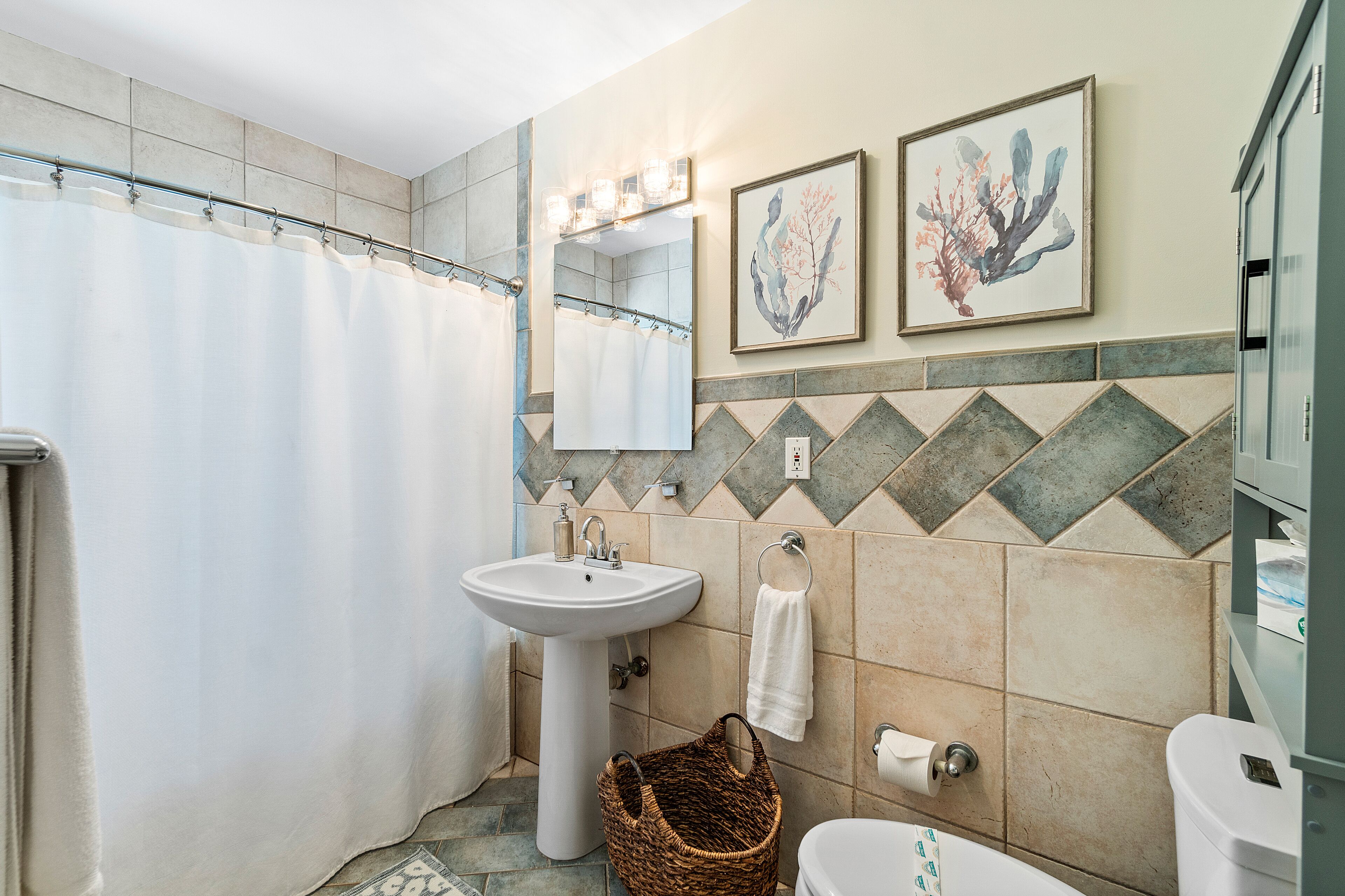 A bathroom with a shower curtain, sink, toilet, and two framed artworks on the wall.