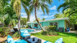 A poolside area with wicker furniture and a small table, surrounded by lush greenery and palm trees. - BY THE SEA VACATION VILLAS 3 BED POOL/ BEACH AT END OF STREET (Lauderdale-by-the-Sea)