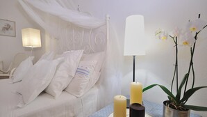 Una cama bien hecha con sábanas y almohadas blancas, una mesita de noche con una lámpara, una orquídea en maceta y dos velas.