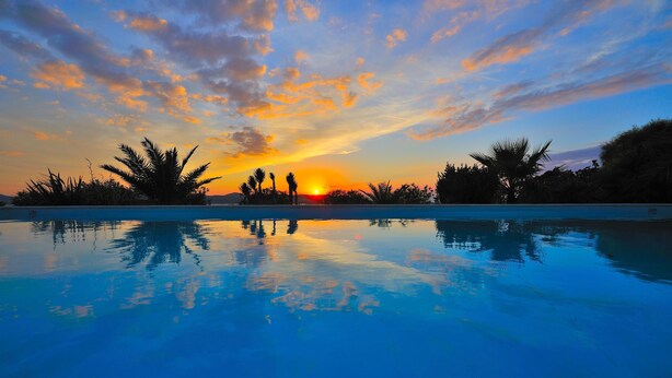 Una piscina refleja una puesta de sol, palmeras y un cielo despejado.