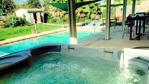 A hot tub area with a pool in the background, surrounded by outdoor furniture and greenery.