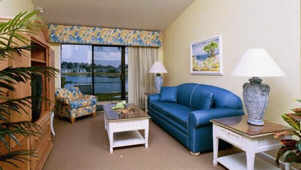 Ein Zimmer mit einem blauen Sofa, einem kleinen weißen Tisch, einem gemusterten Sessel, einem großen Fenster mit Blick auf ein Gewässer und einem Bild an der Wand.