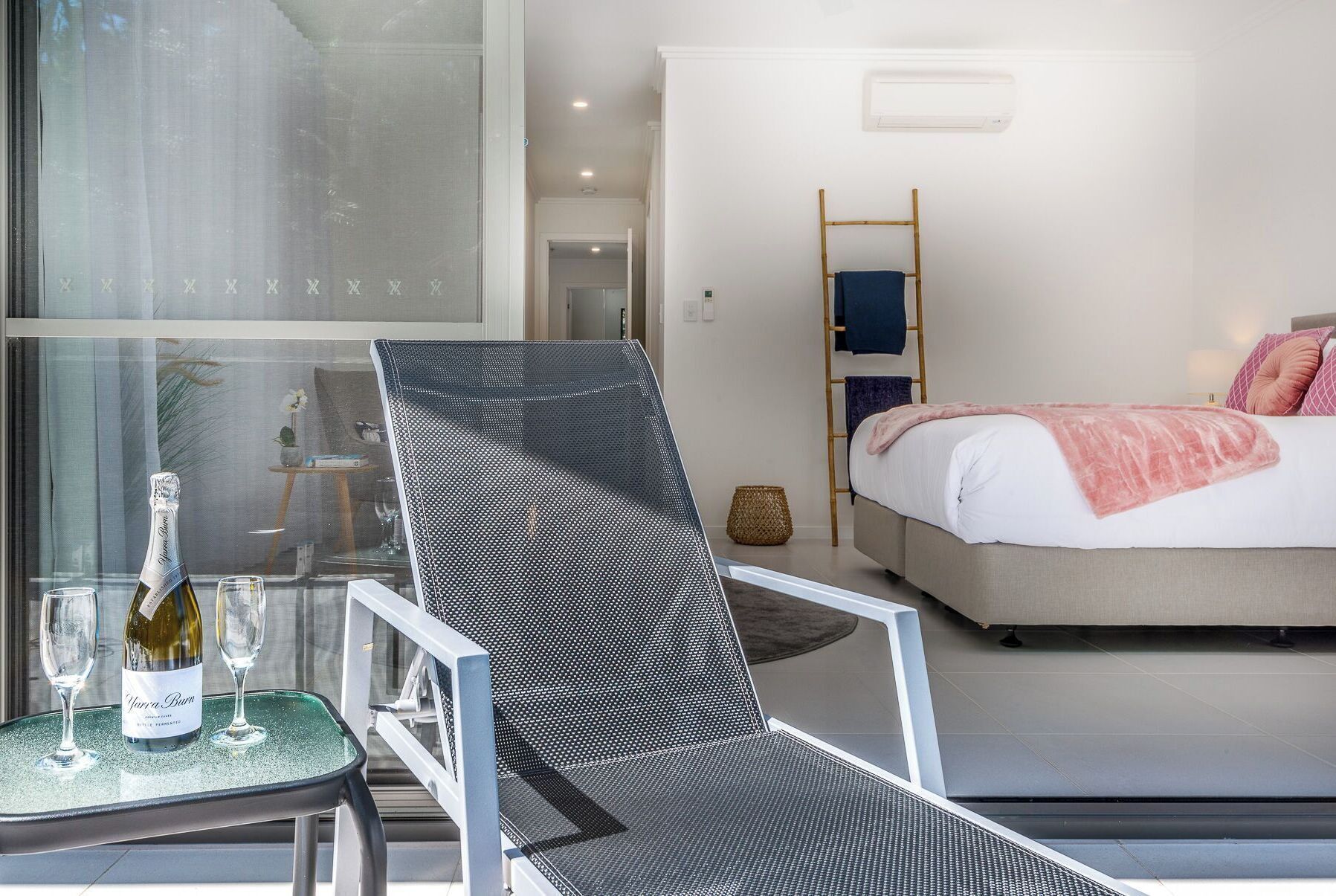 Une chambre d’hôtel moderne avec un grand lit, un coin salon avec un fauteuil et une table en verre sur laquelle se trouvent une bouteille et des verres.