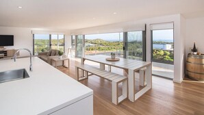 Eine moderne Küche mit einer großen Kochinsel, einem Essbereich mit einem Holztisch und Bänken sowie großen Schiebeglasüren, die einen Blick auf die Landschaft bieten.