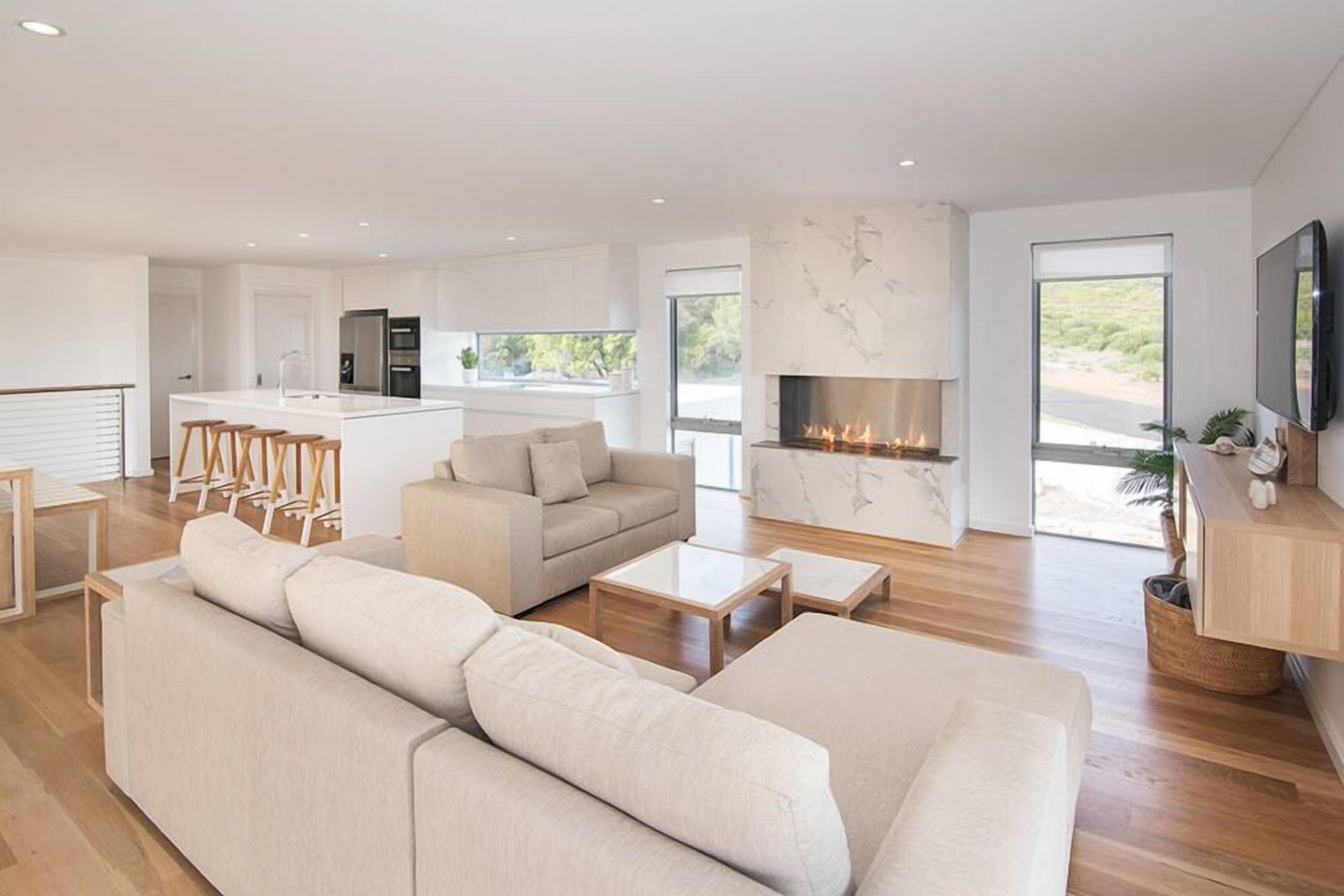 A modern living room with a large beige sectional sofa, a fireplace, and a kitchen area with white cabinets and a marble backsplash.