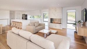 Ein modernes Wohnzimmer mit einem großen, beigefarbenen Ecksofa, einem Kamin und einer Küchenzeile mit weißen Schränken und einer Marmor-Spritzschutzwand.