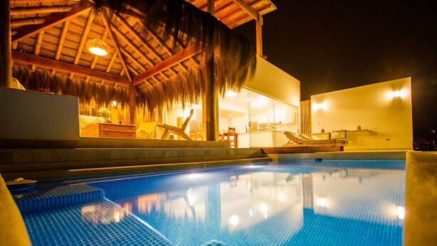 Una piscina con superficie de agua azul cristalina, una estructura de techo de paja y un edificio moderno con grandes ventanales.