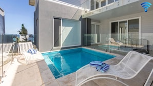 A modern outdoor pool area with clear blue water, white lounge chairs, and a glass-enclosed balcony.