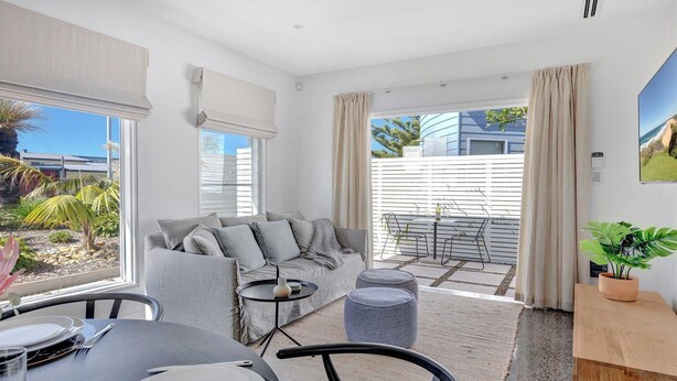 A modern living room with a grey sofa, a dining table, and a small balcony with a view of the sea.