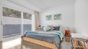 A bedroom with a large sliding glass door, a bed with a grey and blue blanket, two framed plant artworks on the wall, and a small wooden stool with a white towel.