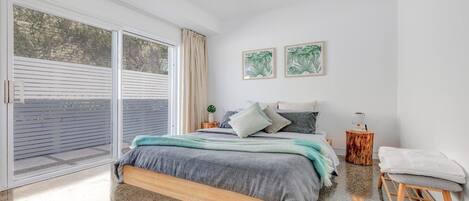 Un dormitorio con un gran ventanal corredera, una cama con manta gris y azul, dos obras de arte con plantas enmarcadas en la pared, y un taburete de madera con una toalla blanca.