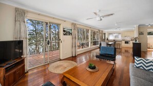 A spacious living area with a large TV, wooden coffee table, and a sofa. There is a balcony with glass doors and a view of trees.