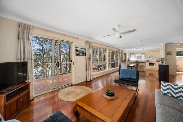 A spacious living area with a large TV, wooden coffee table, and a sofa. There is a balcony with glass doors and a view of trees.