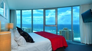 A hotel room with a large bed, a red blanket, a TV on the wall, and a view of the ocean and buildings. - Wake Up In Paradise, Luxury Surfers Paradise Apt (Surfers Paradise)