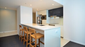 A modern kitchen with white countertops, a built-in oven, and a microwave. There are three wooden bar stools and a white refrigerator. - Wake Up In Paradise, Luxury Surfers Paradise Apt (Surfers Paradise)