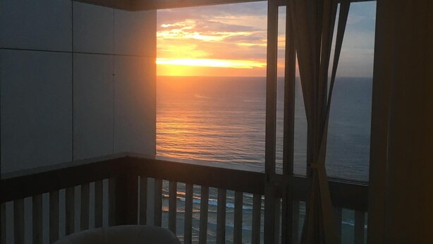 Een kamer met uitzicht op de ondergaande zon boven de oceaan, een balkon met een hekwerk en twee stoelen.