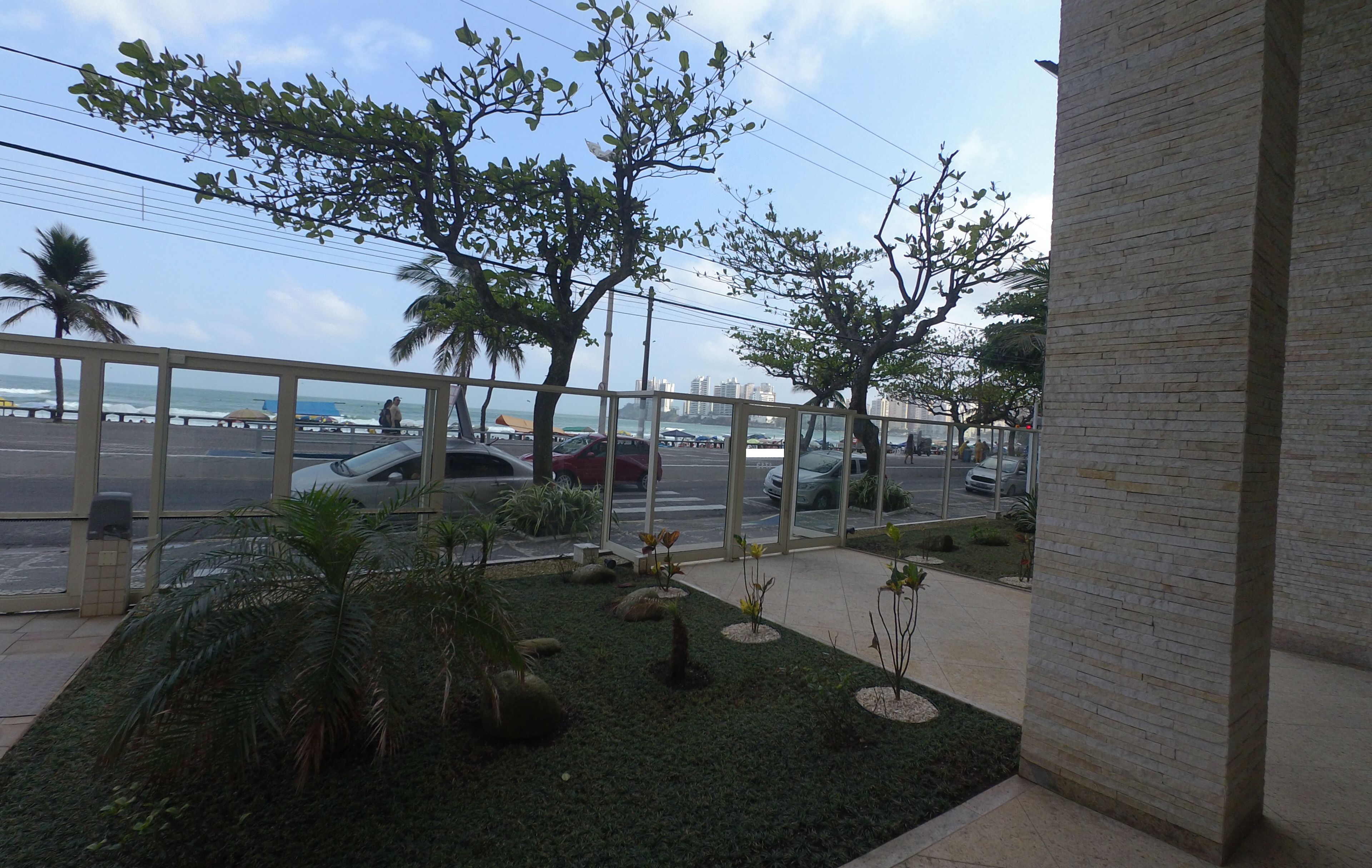 Vista de uma varanda com vista para um estacionamento, palmeiras e uma praia ao fundo.