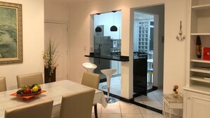 Een moderne eetruimte met een tafel voor twee personen, een keuken zichtbaar door een open doorgang, en een witte wandplank met decoratieve objecten.