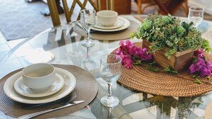 Una mesa redonda de vidrio preparada con platos y cuencos blancos, cubiertos y un mantel tejido. Un macetero de madera con follaje verde y flores rosas se encuentra sobre la mesa.