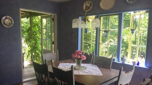 Een eethoek met een houten tafel, stoelen en een bloevas. Op de muur hangen decoratieve borden en er is een raam met uitzicht op groen.