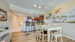 Una cocina amplia con armarios de madera, un comedor con mesa y sillas blancas, y un mural con una escena tropical en la pared.