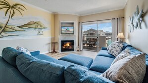 Una sala de estar con chimenea, televisor montado encima y ventilador de techo, con vistas al exterior a través de un ventanal.