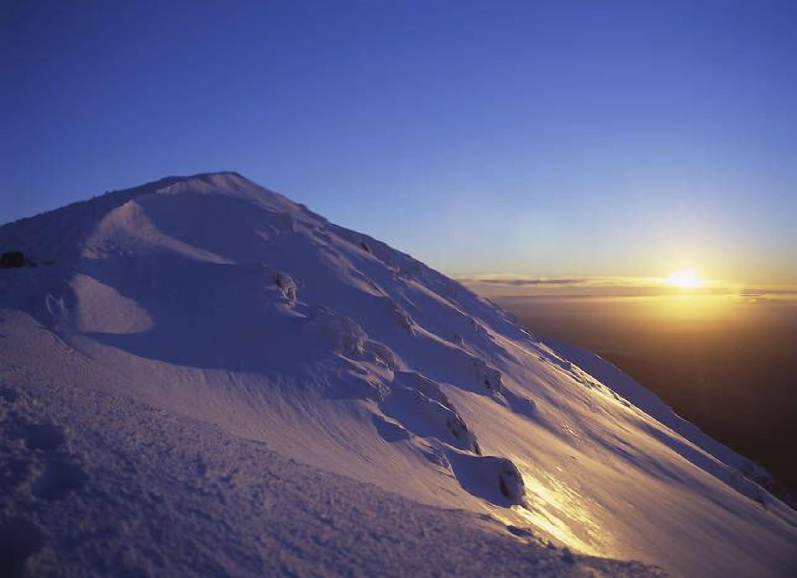 A snow-covered mountain slope with a clear sky and the sun setting in the background.
