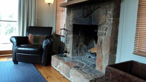 A living room with a stone fireplace, a leather armchair, a floor lamp, and a view of the outdoors through a window with blinds. - Tikkandi – Cozy Luxe Getaway in Victoria’s High Country (Merrijig)
