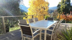 A deck with a glass-top table and six chairs, surrounded by a wooden deck and a view of trees and mountains. - Tikkandi – Cozy Luxe Getaway in Victoria’s High Country (Merrijig)
