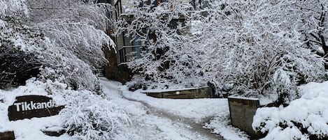 Experience the charm of Tikkand, a traditional house surrounded by snow-covered trees and a pathway leading to its doorstep.