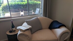 Ein beiges Sofa mit einem Buch und einer Weinflasche auf einem kleinen runden Tisch neben einem Fenster.