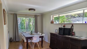 Ein Essbereich mit Tisch und Stühlen, einer dunklen Holzkommode und einem großen Fenster mit Grünpflanzen außerhalb.