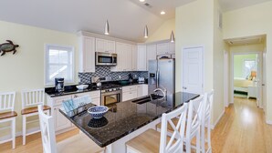 A modern kitchen with white cabinets, a black granite countertop, and stainless steel appliances. There are white chairs around the island and a view into the adjacent living area. - Calypso by Seaside Vacations & Sales (Greenbackville)