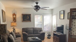 A living room with a sofa, ottoman, and a television. There is a ceiling fan, a clock, and a lamp.