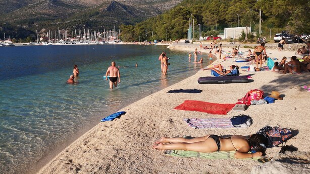 Una spiaggia con acqua cristallina, persone che prendono il sole e imbarcazioni sullo sfondo.