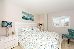 A bedroom with a bed, a nightstand, a lamp, a chair, and artwork on the walls. - Beachcomber unit 403 (Longboat Key)