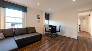 A modern living room with a dark grey sofa, a dining table with chairs, and a wooden floor. - Stunning Hyde Park Apartment (SS3) (London)